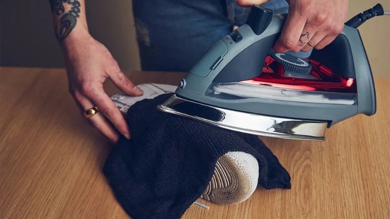 Một người đang cẩn thận dùng bàn ủi hơi nước để làm phẳng các vết nhăn trên bề mặt giày sneaker da, có lót vải bảo vệ